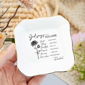 Può includere: Un piatto portagioie in ceramica bianca con bordo dorato. Il piatto presenta il testo "Dio dice CHE SEI" e parole ispiratrici con riferimenti biblici. È presente anche un'illustrazione floreale.
