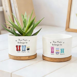 May include: Two white ceramic planters with bamboo bases. One planter holds a green succulent. Both planters have the text "This Mum belongs to" and feature colorful boot illustrations, names, and a paw print. A framed picture and books are in the background.