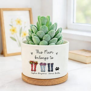 Peut inclure: Un pot de fleurs en céramique blanche avec une plante succulente. Le pot porte l'inscription "This Mum belongs to" et des illustrations de bottes avec des fleurs et une empreinte de patte. Les noms Sophia, Benjamin, James et Bella sont en dessous.