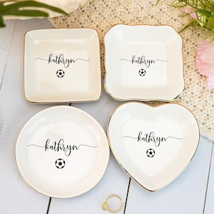 May include: Four white ceramic trinket dishes in square, scalloped square, round, and heart shapes. Each dish has a gold rim, the name "kathryn" in script, and a soccer ball graphic. A gold ring is in the foreground.
