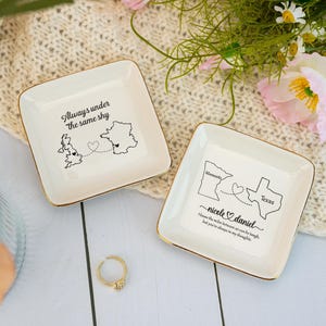 Op de afbeelding: Twee vierkante, crèmekleurige keramische sieradenbakjes met gouden randen. Eén bakje toont een kaart van het VK en Frankrijk met de tekst "Always under the same sky". Het andere bakje toont kaarten van Minnesota en Texas met de namen "nicole & daniel".