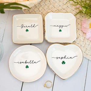 May include: Four white ceramic trinket dishes with gold rims. Each dish has a name in black script and a green shamrock. The dishes are square, round, and heart-shaped.