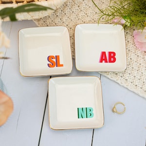 Może przedstawiać: Trzy kwadratowe białe ceramiczne miseczki na drobiazgi ze złotymi obwódkami. Każda miseczka posiada dwuliterowy monogram wykonany wyrazistymi, kolorowymi literami blokowymi: SL w kolorze pomarańczowym i fioletowym, AB w czerwonym i różowym oraz NB w zieleni.