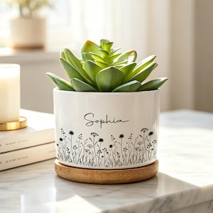May include: A white ceramic cylindrical planter featuring a black line art illustration of wildflowers around the base and the name Sophia printed in black script. The planter sits on a circular bamboo saucer and contains a green succulent plant.