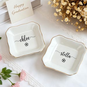 Peut inclure: Deux petits plats à bijoux en céramique blanche avec des bords dorés, chacun portant un nom et un ballon de football. L'un porte l'inscription "chloe", l'autre "stella". Une carte avec "Happy Graduation" est visible.