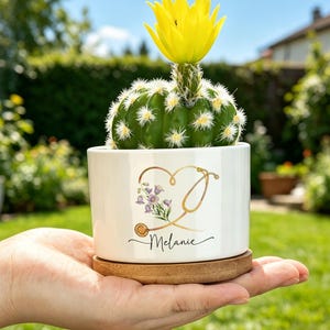 May include: A small green cactus with a bright yellow flower in a white ceramic planter. The planter features a gold stethoscope graphic forming a heart shape with purple flowers and the name Melanie printed in black script on a bamboo base.