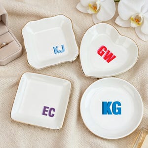 Könnte beinhalten: Vier weiße Keramikschalen für Schmuck mit goldfarbenen Rändern und personalisierten Initialen in verschiedenen Farben. Eine quadratische Schale mit 'KJ', eine herzförmige Schale mit 'GW', eine quadratische Schale mit 'EC' und eine runde Schale mit 'KG'.
