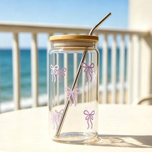 Könnte beinhalten: Ein klares Glas mit einem Bambusdeckel und einem Edelstahlstrohhalm. Das Glas ist mit hellvioletten Schleifendesigns verziert. Der Becher steht auf einem weißen Tisch mit Blick auf das Meer im Hintergrund.