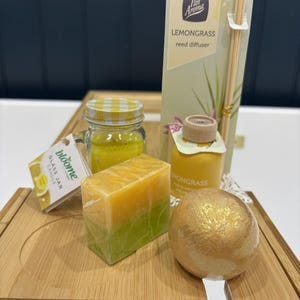 Op de afbeelding: Een verzameling bad- en spaproducten, waaronder een gele glazen potkaars, een citroengras rietverspreider, een stuk zeep en een gouden badbruisbal. De items zijn gerangschikt op een houten dienblad.