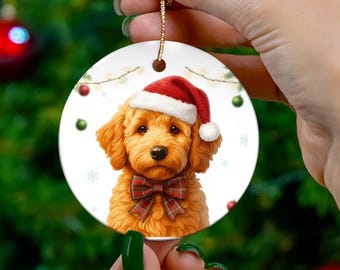 May include: A round Christmas ornament featuring a golden doodle wearing a Santa hat and a plaid bow tie. The ornament has a white background with snowflakes and Christmas lights. A gold string is attached for hanging.