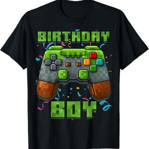 May include: Black t-shirt featuring a pixelated video game controller in green, gray, orange, and brown. The words "BIRTHDAY BOY" are printed above and below the controller in green pixelated letters. Colorful confetti surrounds the design.