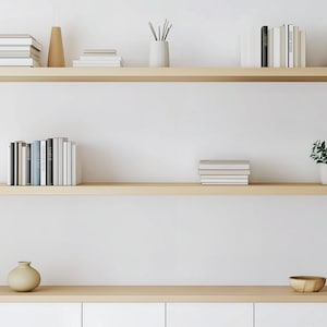 May include: A minimalist interior design featuring three wooden shelves against a white wall. The shelves display books, small potted plants, vases, and decorative objects. The overall aesthetic is clean and modern.