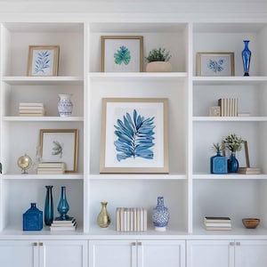 May include: A white built-in bookshelf with various decorative items. The shelves display framed botanical prints, blue vases, stacks of books, and a small gold globe. The color scheme is primarily white, gold, and shades of blue.