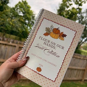 May include: A spiral-bound notebook with a cream cover featuring a brown polka dot pattern and a scalloped red border. The cover displays an illustration of pumpkins and leaves with the text: Flour on our hands: Fall Edition, Jade Cline Lori Sanders, 2026.