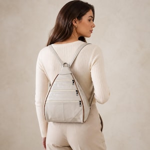 May include: A light gray triangular backpack featuring three horizontal zippered compartments on the front, thin shoulder straps, and brown piping trim along the edges. The bag has a structured shape with a smooth finish and dark metal zipper pulls.