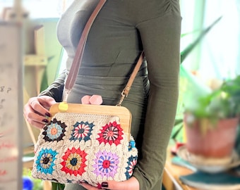 Puede incluir: Un bolso de crochet beige con marco de madera y correa de cuero marrón. El bolso presenta patrones florales coloridos en rojo, azul, amarillo y morado. El bolso lo sostiene una persona que lleva un vestido largo verde oliva.