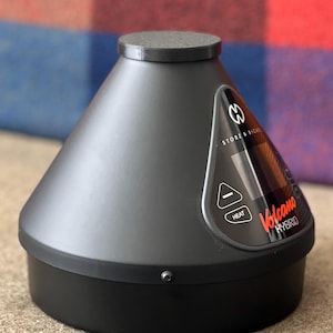 May include: A black and gray Volcano Hybrid vaporizer. The device has a conical shape with a rounded base and a flat top. The control panel displays the word "HEAT" and the brand name "Volcano Hybrid" in red.