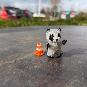 Puede incluir: Una figura de panda hecha a mano con cuentas negras y blancas, junto a un pequeño cono de cuentas naranja y blanco. El panda tiene ojos, orejas y extremidades de cuentas negras. El cono tiene un diseño de cono de tráfico.