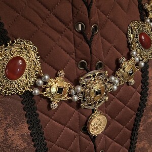 May include: A gold necklace with a large pendant and smaller charms. The necklace is made of metal and has a pearl-like bead detail. The pendant features a black stone and intricate designs. The necklace is on a brown quilted fabric background.