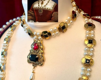May include: A pearl necklace with gold accents and jewel-toned embellishments. The necklace features a pendant with a red and black stone, and is displayed on a mannequin. A portrait of a woman in historical attire is in the background.