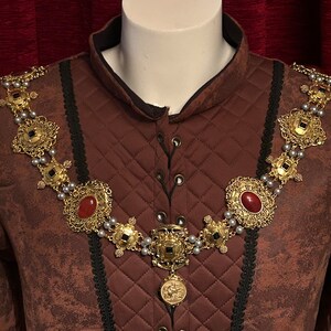 May include: A gold chain necklace with red and black gemstones. The chain is made of intricate metalwork and features a pendant with a design of a lion.