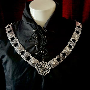 May include: A silver chain necklace with a decorative pendant featuring a stylized design. The chain is made up of square links with ornate details and features a central pendant with a detailed design.