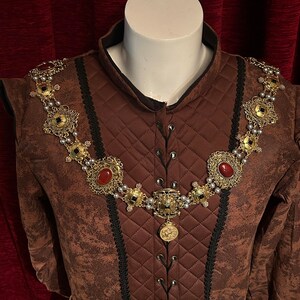 May include: A brown quilted jacket with black trim and a gold chain necklace with red and black gemstones. The necklace has a large pendant with a gold design.