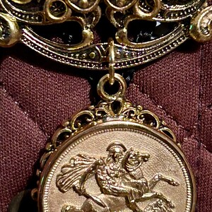 May include: A gold-toned pendant with a detailed design featuring a knight riding a horse. The pendant is attached to a black cord.