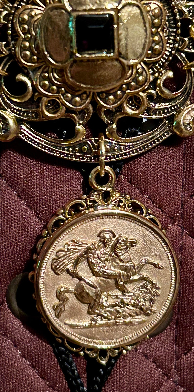May include: A gold-toned pendant with a detailed design featuring a knight riding a horse. The pendant is attached to a black cord.