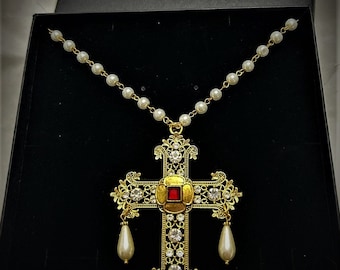 May include: A gold cross necklace with a red jewel in the center and pearl accents. The necklace is in a black box with the text "The Enchanted Tudor Rose".