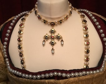 May include: A gold-toned necklace with a cross pendant, adorned with red and black gemstones and pearls, displayed on a mannequin. The necklace is part of a historical costume, with a gold patterned bodice and a pearl-trimmed neckline.