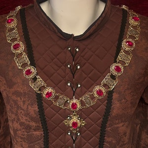 May include: A gold chain necklace with red gemstone accents. The chain is made of interlocking circles and squares. The necklace has a pendant in the shape of a cross with a red gemstone in the centre.