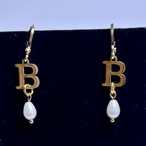 May include: A pair of gold-toned hoop earrings featuring a dangling capital letter B charm and a small, white, teardrop-shaped faux pearl pendant suspended from the bottom of each letter. The earrings have a simple, polished metal finish.