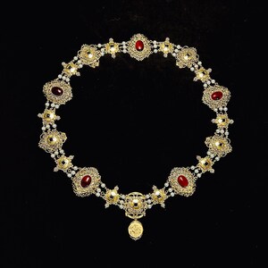 May include: A gold necklace with a pendant featuring a small image of a person. The necklace is adorned with red gemstones and white pearls.