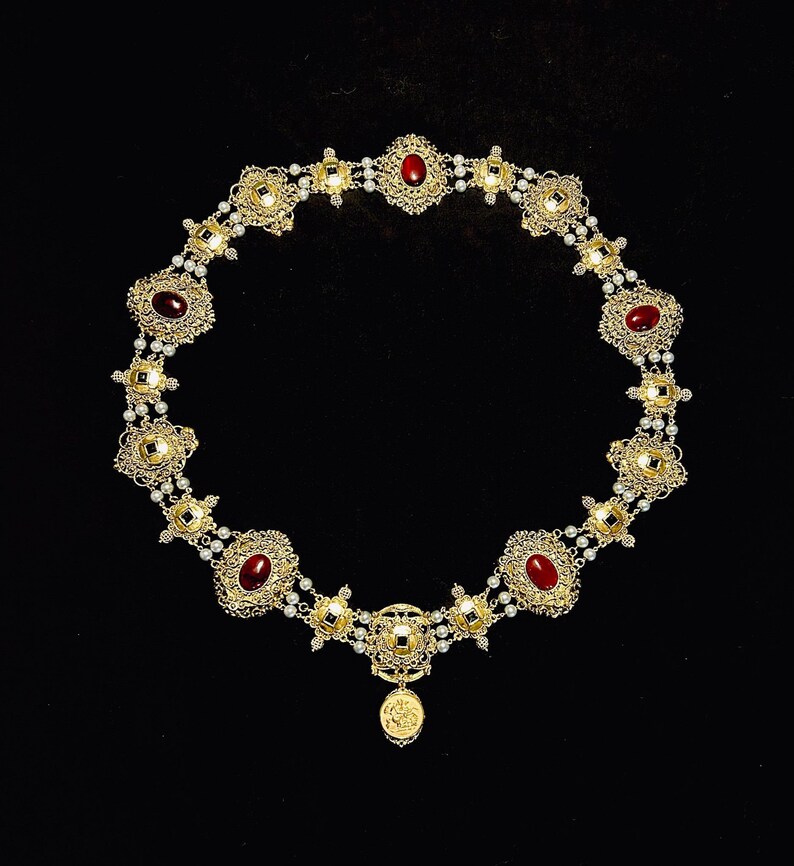 May include: A gold necklace with a pendant featuring a small image of a person. The necklace is adorned with red gemstones and white pearls.