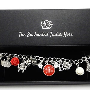 May include: A silver charm bracelet with a variety of charms, including a red rose, a crown, a dice, a book, a cross, a horse, a white rose, and a charm that says "The More The Merrier". The bracelet is in a black box with the words "The Enchanted Tudor Rose" on the lid.