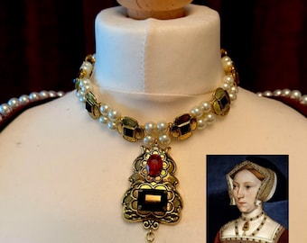 May include: A gold-toned necklace with faux pearls and jewel accents. The necklace features a pendant with a large red and black jewel, and a dangling pearl. A portrait of a woman wearing a similar necklace is in the corner.