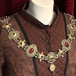 May include: A brown quilted garment with a gold and silver chain necklace featuring red gemstone accents. The necklace has a pendant with a detailed design.