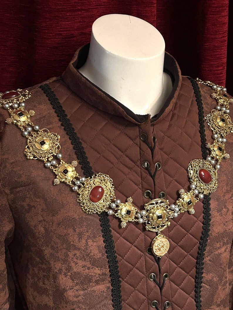 May include: A brown quilted garment with a gold and silver chain necklace featuring red gemstone accents. The necklace has a pendant with a detailed design.