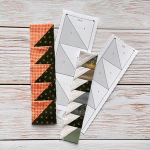 May include: Two fabric strips with triangle patterns in orange and green, and a second strip with a variety of neutral colors. Two paper templates with triangle shapes and measurements of 2.5 inches and 2 inches are also present.