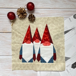 May include: A fabric square featuring three gnome figures with red hats and white beards. The gnomes are set against a beige background with a subtle floral pattern. Red ornaments and gold pinecones are placed around the square. Paper templates are also visible.