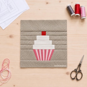 Könnte beinhalten: Ein quadratischer Stoff-Quiltblock mit einem zentralen Cupcake-Motiv. Der Cupcake hat ein weißes Frosting, eine rote Kirsche und eine rosa-weiß gestreifte Basis, alles vor einem beigen Hintergrund mit einem kleinen, dunklen Punktmuster.