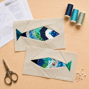 Può includere: Due blocchi di tessuto rettangolari per trapuntatura con un design a pesce composto da pezzi in tonalità blu, verdi e bianche, incorniciati da un bordo beige testurizzato. Il set include tre rocchetti di filo, forbici in metallo e un foglio di istruzioni.