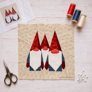 May include: A square fabric quilt block featuring three pieced gnomes with red hats, white beards, and blue patterned bodies on a cream background with small brown sprigs. Includes three spools of thread, sewing scissors, and pins.