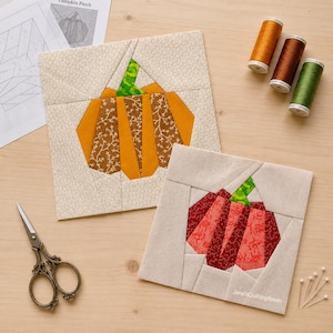 Puede incluir: Dos bloques de tela cuadrados para acolchar con diseños de calabaza. Un bloque muestra una calabaza naranja y marrón, el otro una calabaza roja y granate. Ambos tienen tallos verdes y fondos crema. Incluye tijeras de costura, tres carretes de hilo y patrones de papel.
