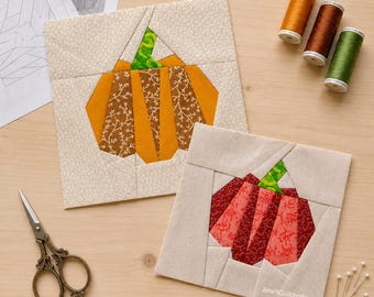 May include: Two square fabric quilt blocks featuring pumpkin designs. One block displays an orange and brown pumpkin, the other a red and maroon pumpkin. Both have green stems and cream backgrounds. Includes sewing scissors, three spools of thread, and paper patterns.