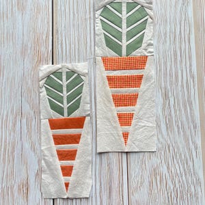 Op de afbeelding: Twee stoffen wortelblokken met groene toppen en oranje lichamen. De worteltoppen hebben een bladvorm met witte accenten. De wortellichamen hebben horizontale oranje strepen. De blokken liggen op een lichte houten achtergrond.