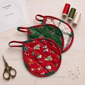 Könnte beinhalten: Zwei runde, gesteppte Topflappen mit Stoffschlaufen. Einer ist rot mit einem Weihnachtsmuster aus Schneemännern und Bäumen, der andere grün mit Beerenmuster. Enthält ein Papierschnittmuster mit der Aufschrift POT HOLDER Cut 2, drei Garnrollen und eine Schere.