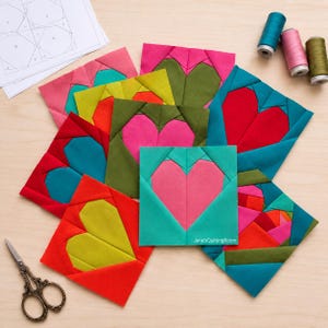 Puede incluir: Una colección de bloques de tela cuadrados para acolchar con diseños de corazones en varios colores, incluyendo rosa, rojo, amarillo, verde y azul cerceta. Los bloques están apilados con un patrón de papel, tres carretes de hilo y tijeras.
