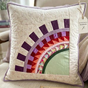 May include: A square quilted pillow featuring a central rainbow arc design composed of purple, red, orange, and green fabric segments. The background is a cream fabric with a subtle floral print. A tag reads Jane'sQuiltingRoom Handmade and 14" QUILTING BLOCK.